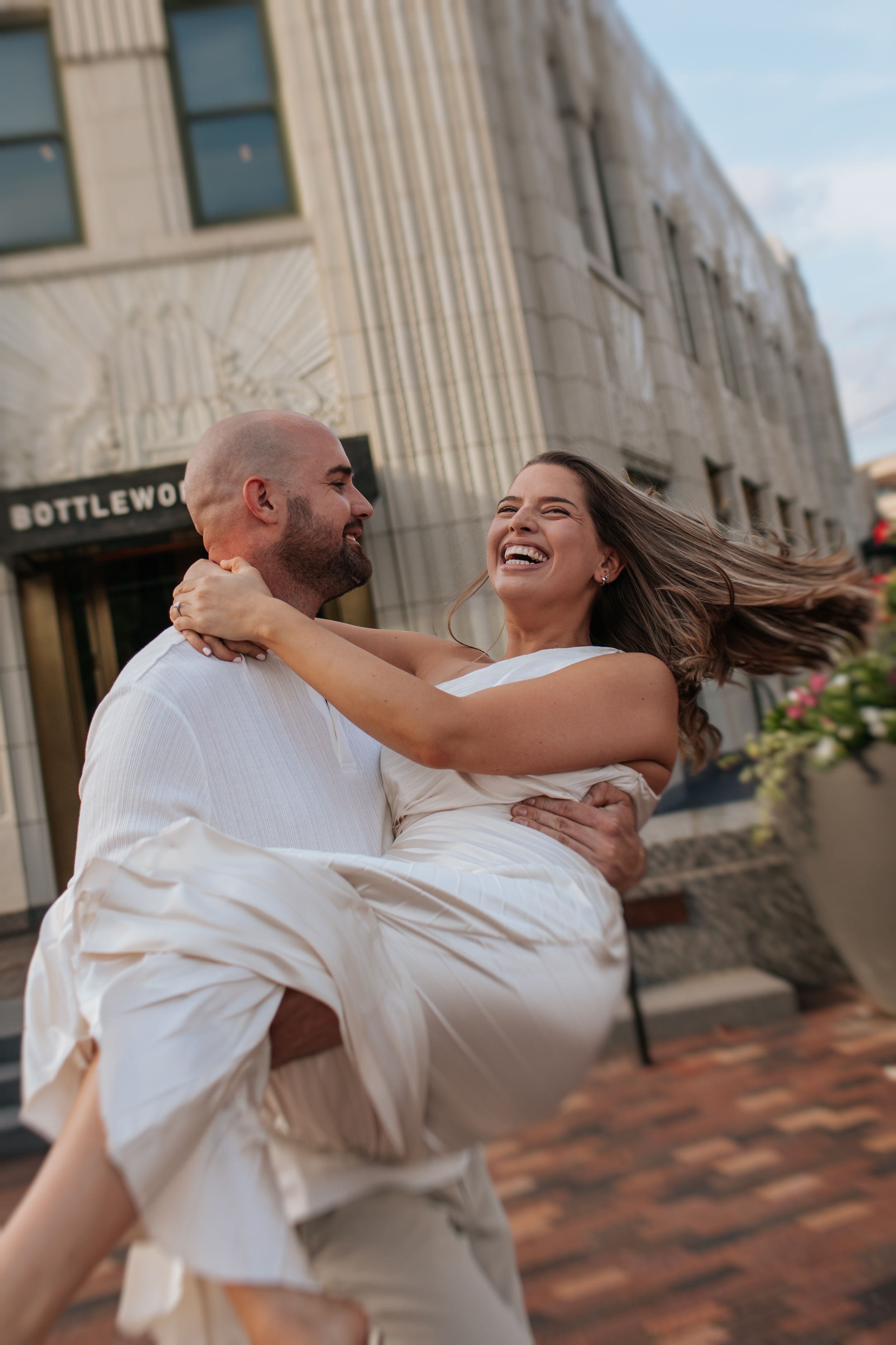 A couple in white clothing spins and dances joyfully together on a brick sidewalk outside a building.