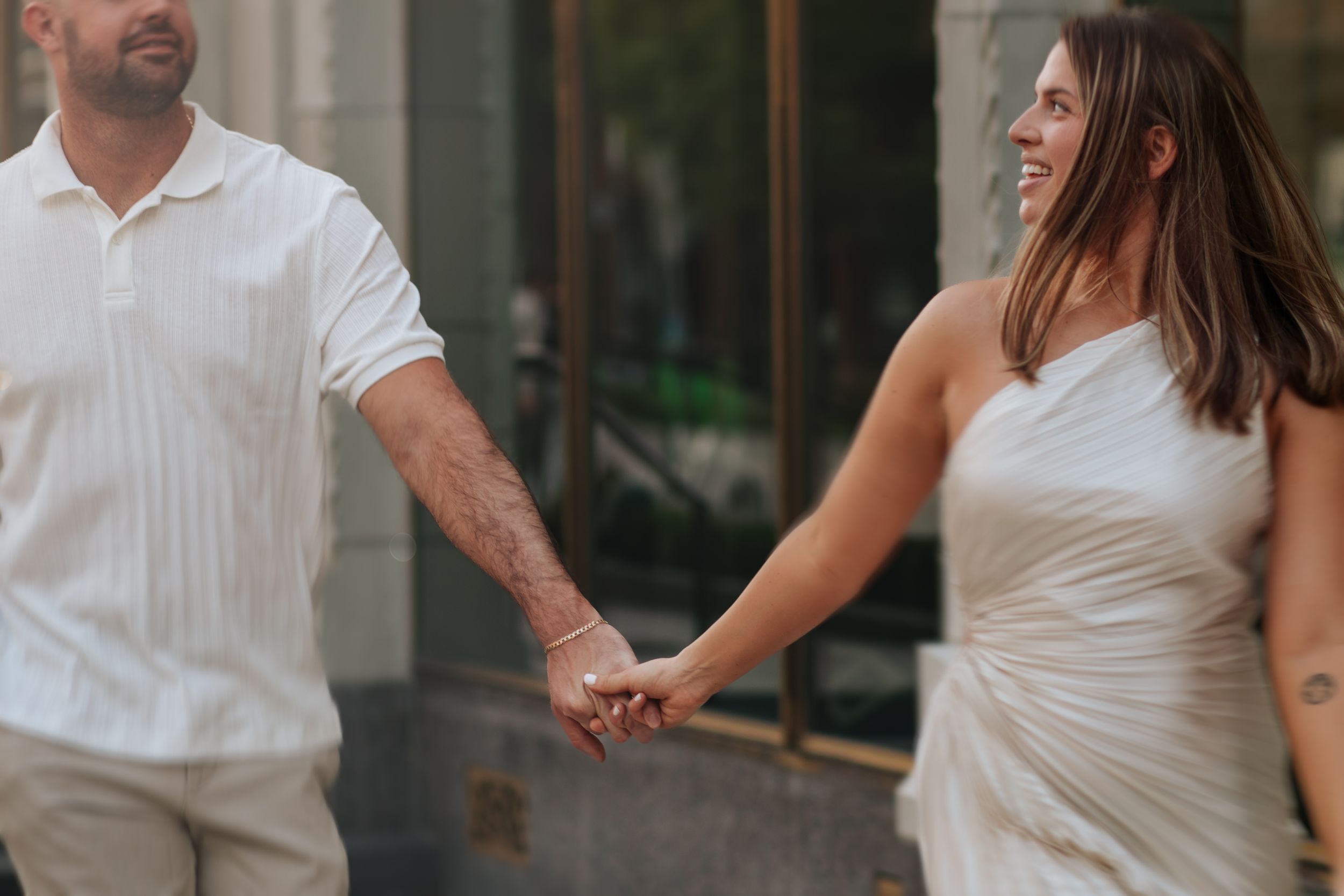 A couple in white attire hold hands while walking together outside a building.