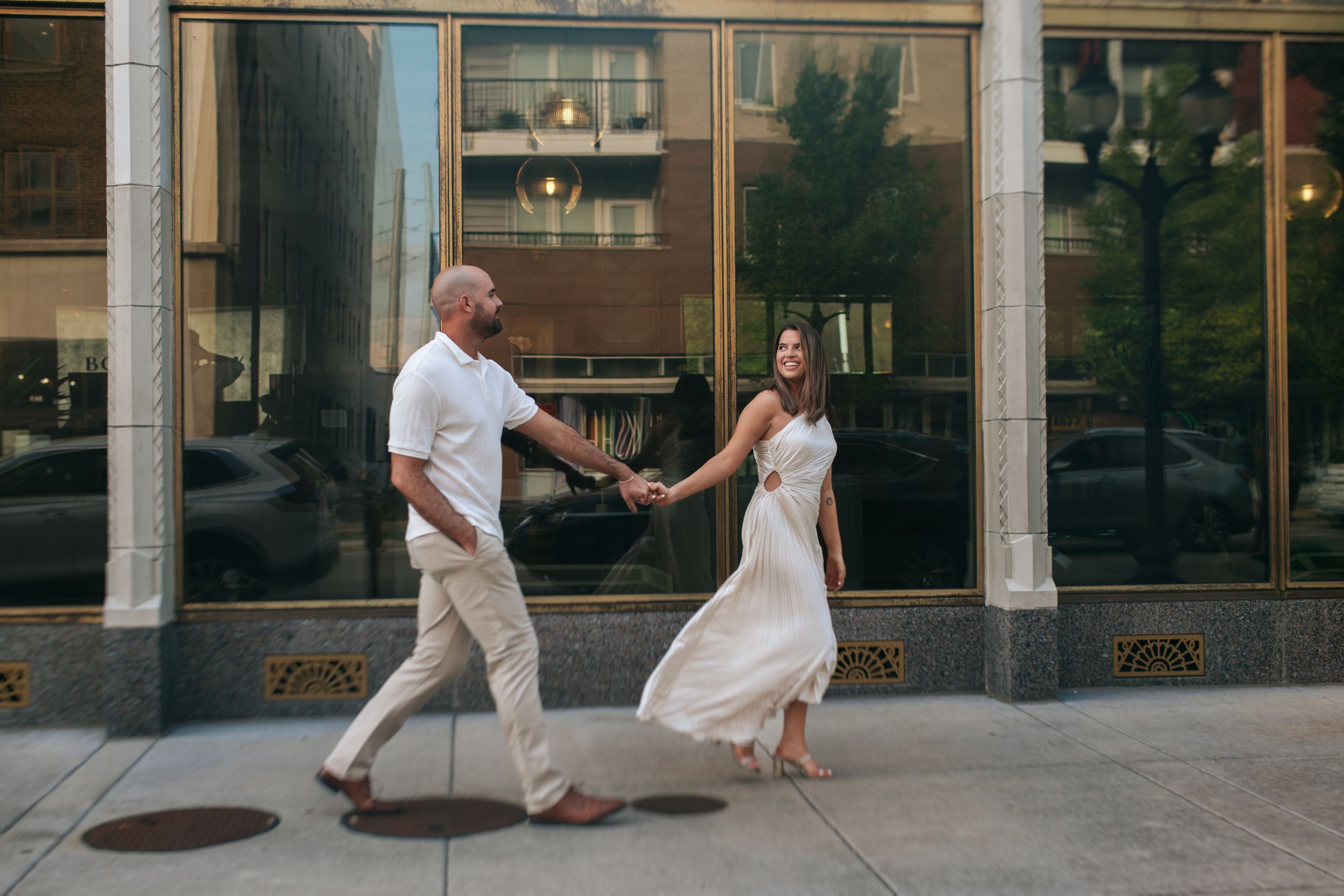 A couple dressed in white shares a playful moment while walking outside an urban building.