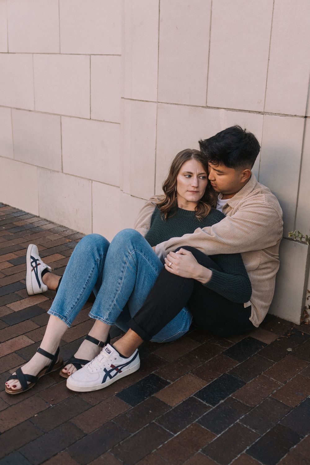 A couple sits intimately together against a white wall on brick pavement wearing casual clothing and sneakers.