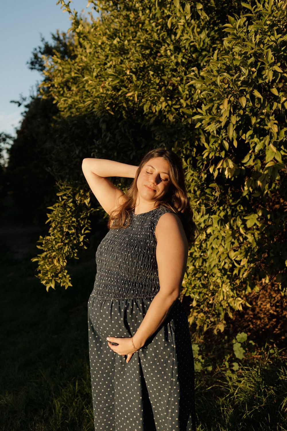 Woman in polka dot dress poses among leafy branches at sunset.