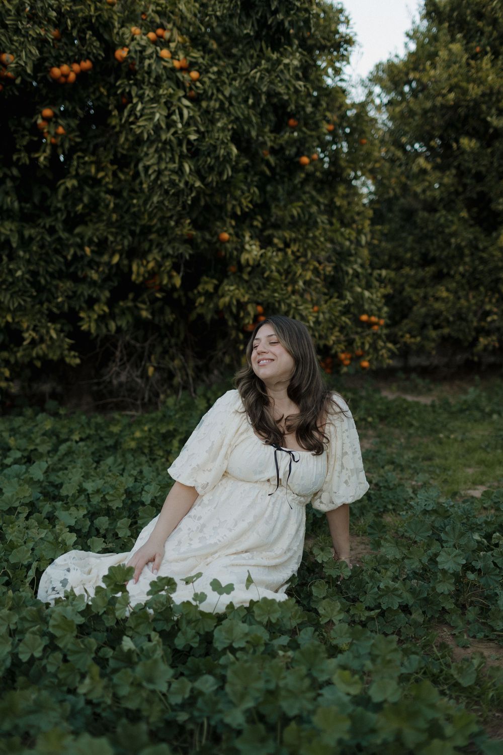 A dreamy maternity photo in an orange grove with someone wearing a vintage-style white dress.