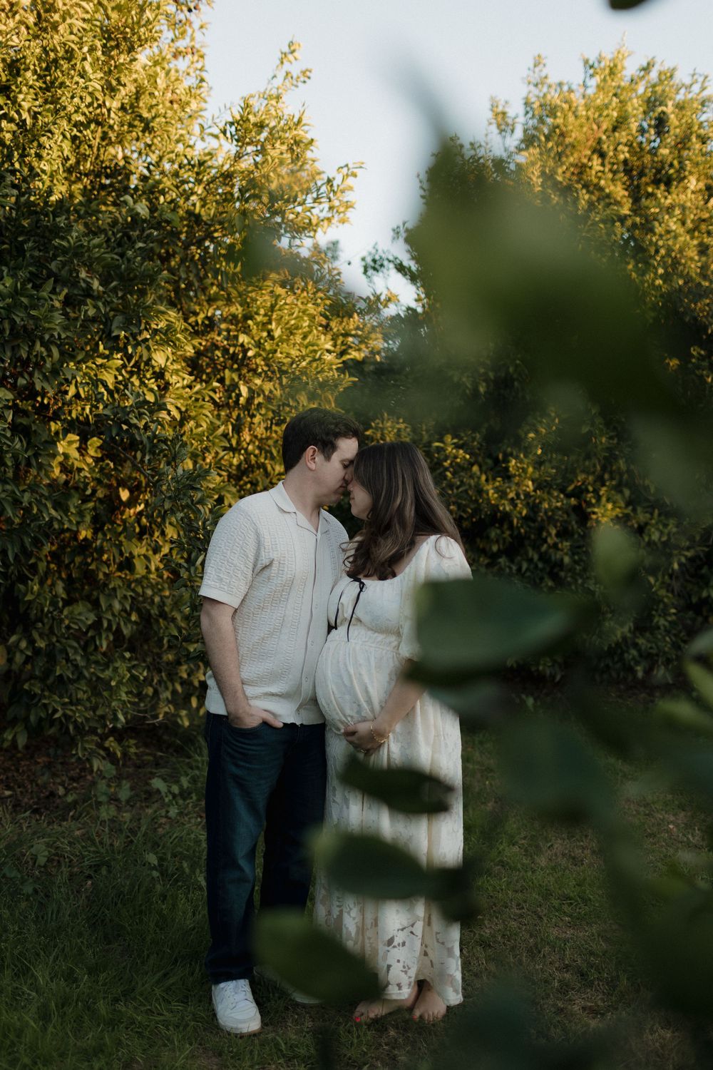 Romantic maternity photo through leaves at sunset in white dress and casual attire.