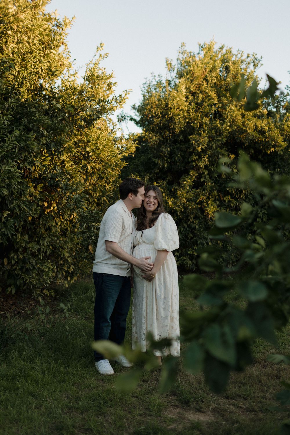 Intimate maternity portrait in an orchard with golden evening light filtering through trees.