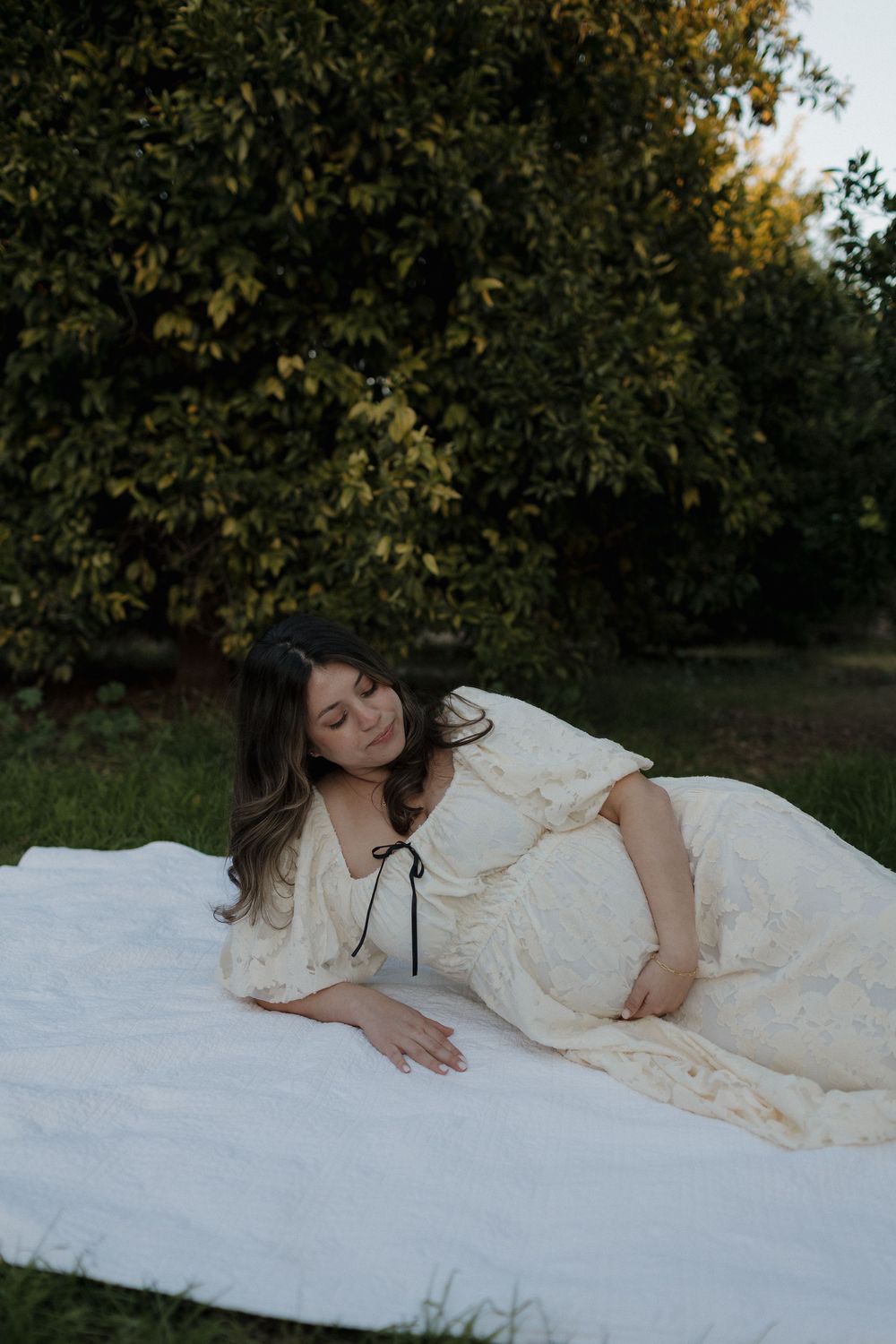 A person in a white lace dress lying gracefully on a white blanket in a garden setting during golden hour.