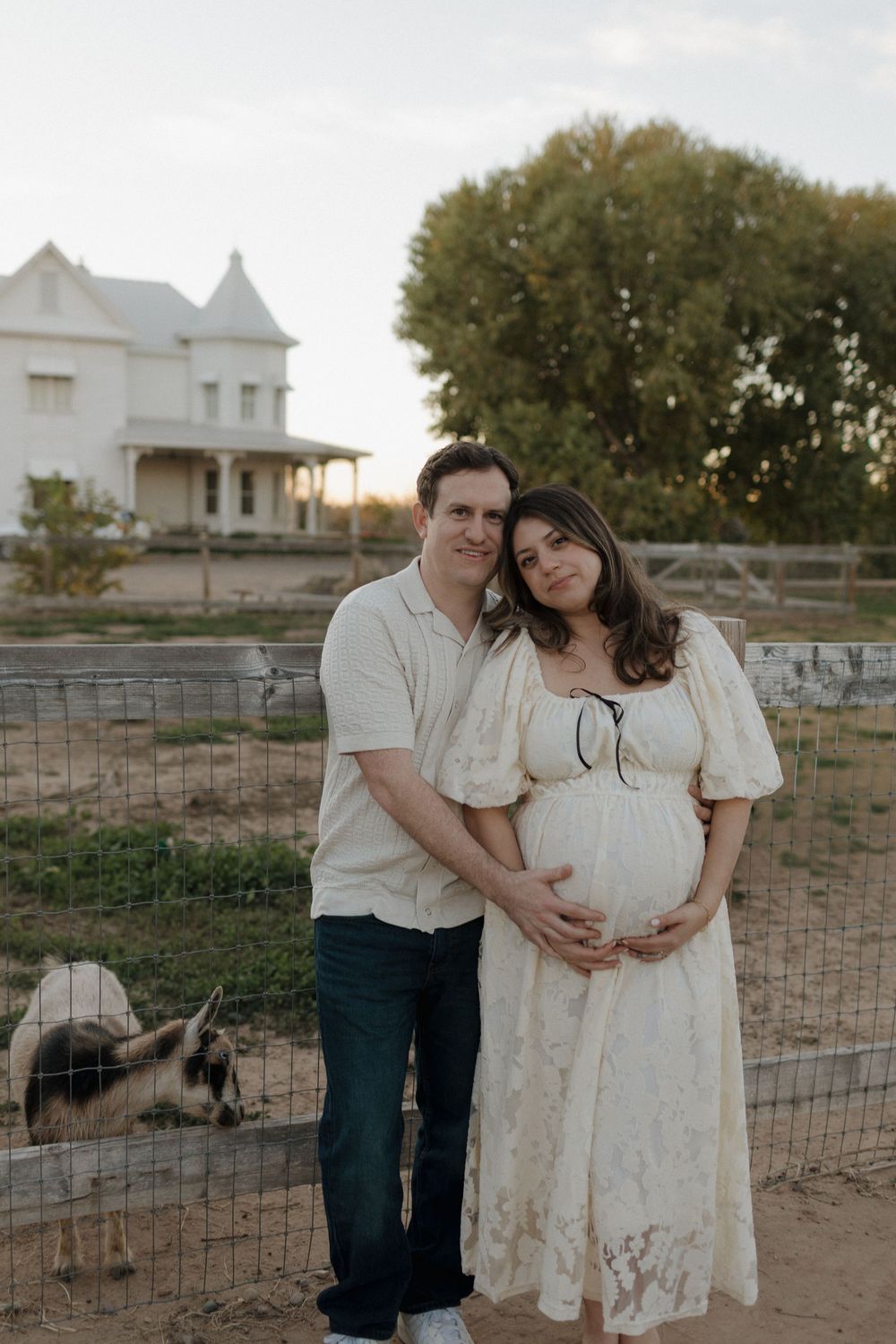A couple embraces during their maternity session near a historic farmhouse at golden hour.