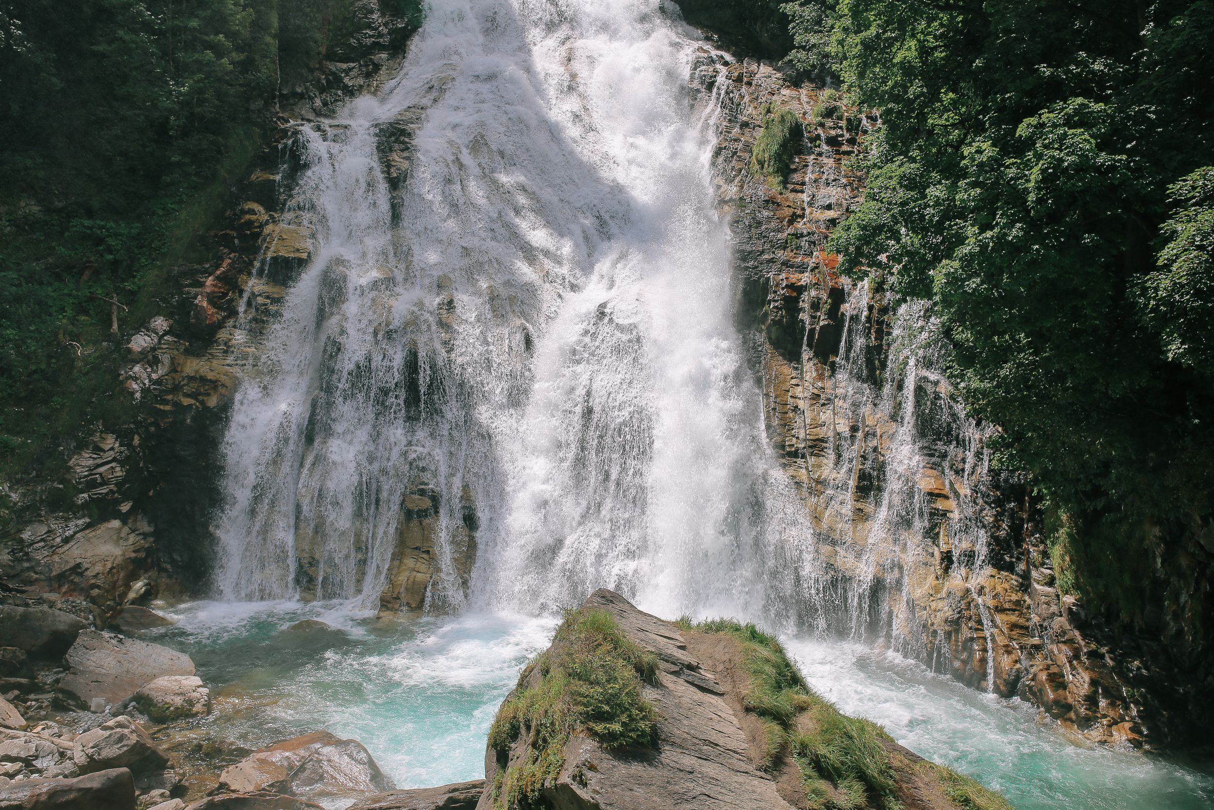 Cascading waterfall flows down rocky cliffs into turquoise pool surrounded by lush forest in scenic mountain landscape.