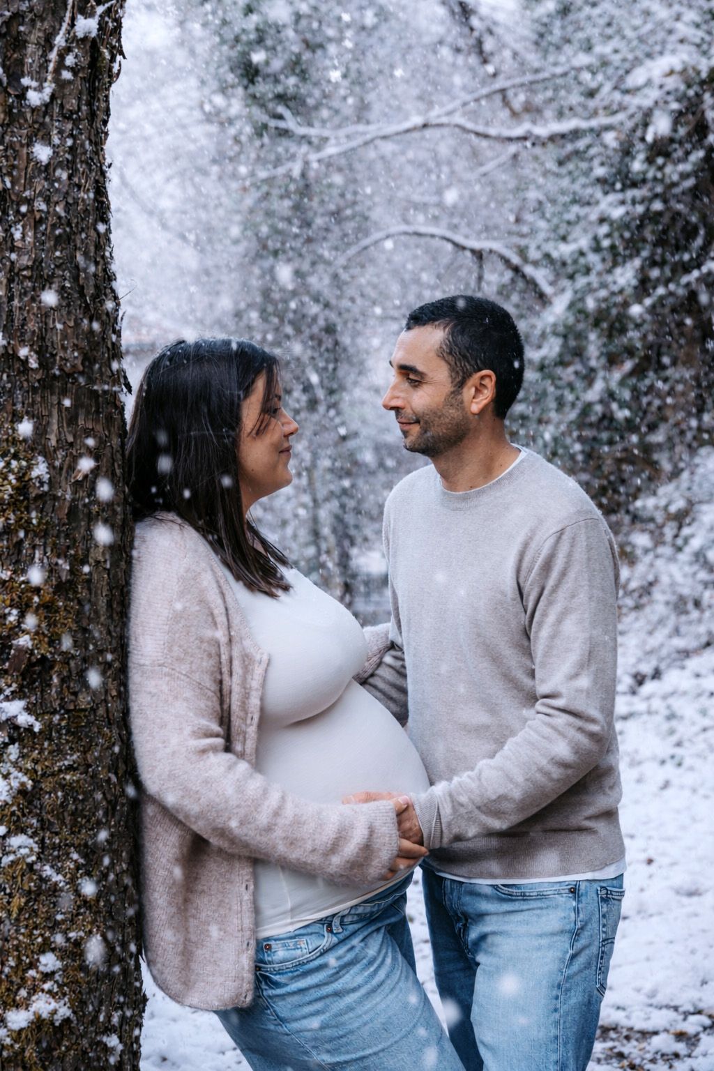 A couple shares tender moments in a snowy winter forest setting while wearing matching light colored sweaters.