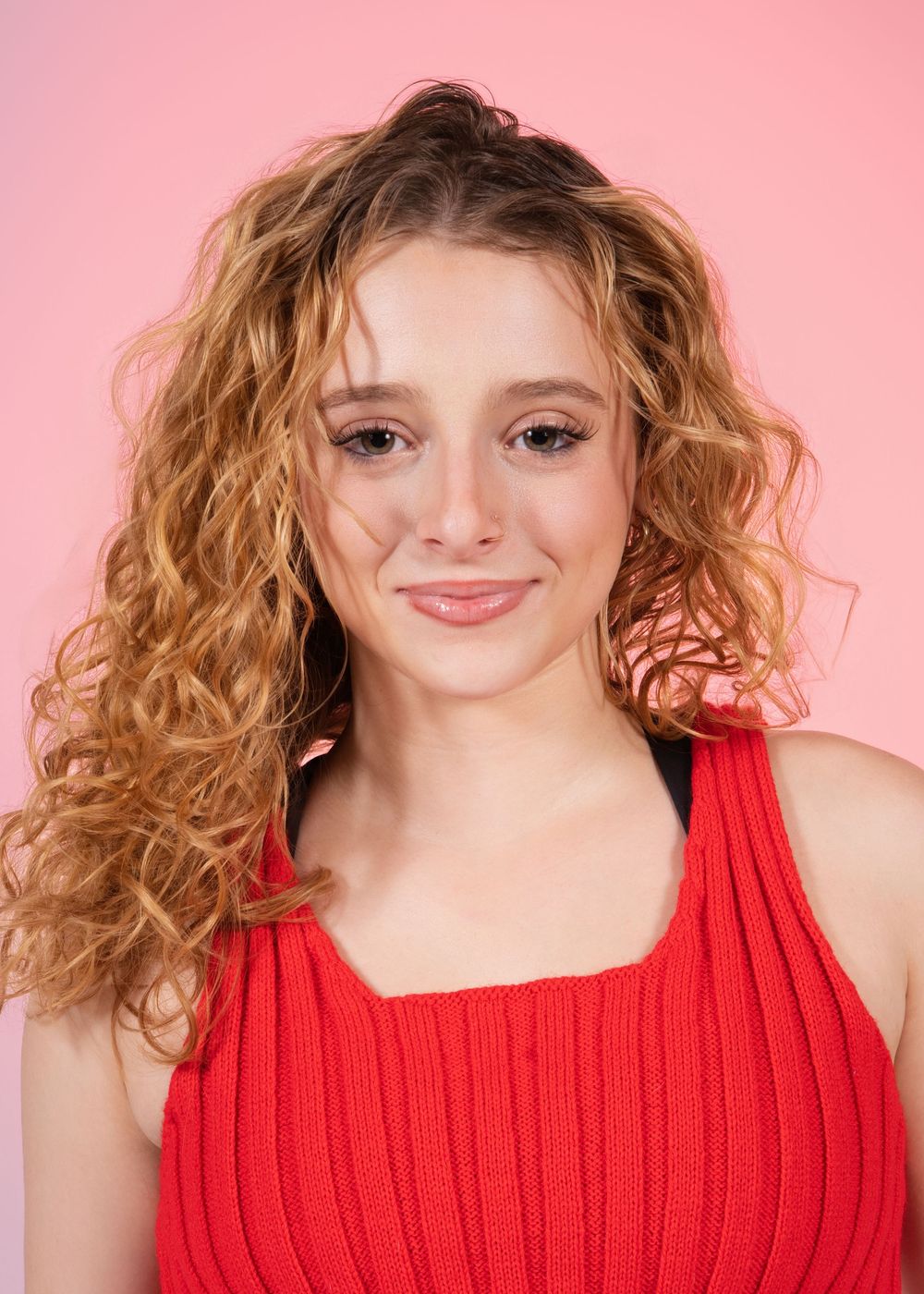 Young person with curly blonde hair wearing a red ribbed tank top poses against a pink background. In studio in Orlando, Florida