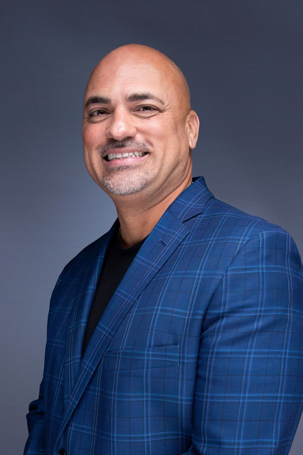 Professional headshot against blue background showing person in plaid blue blazer and black shirt.