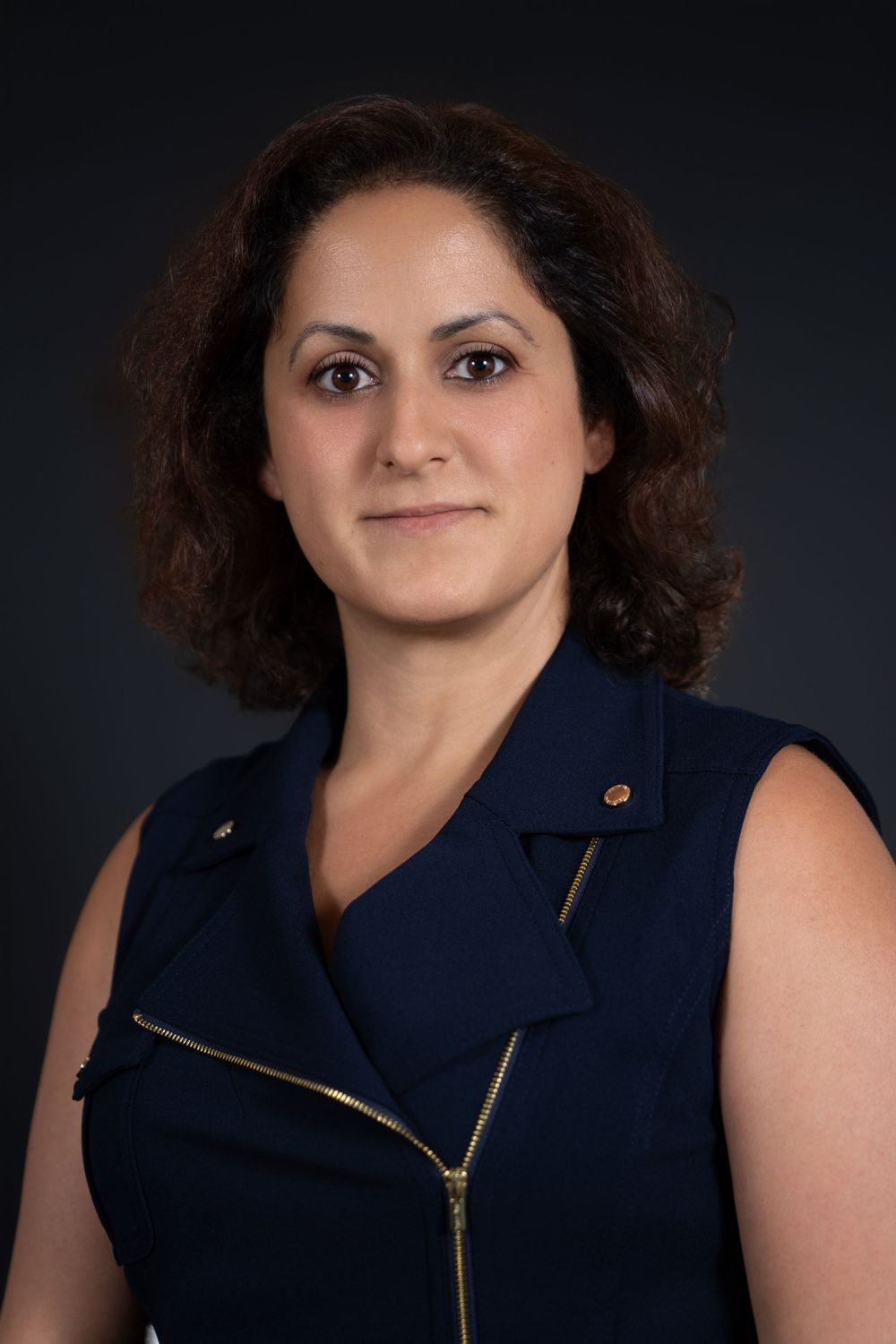 Professional headshot portrait of a person with curly dark hair wearing a navy sleeveless top against a dark background.