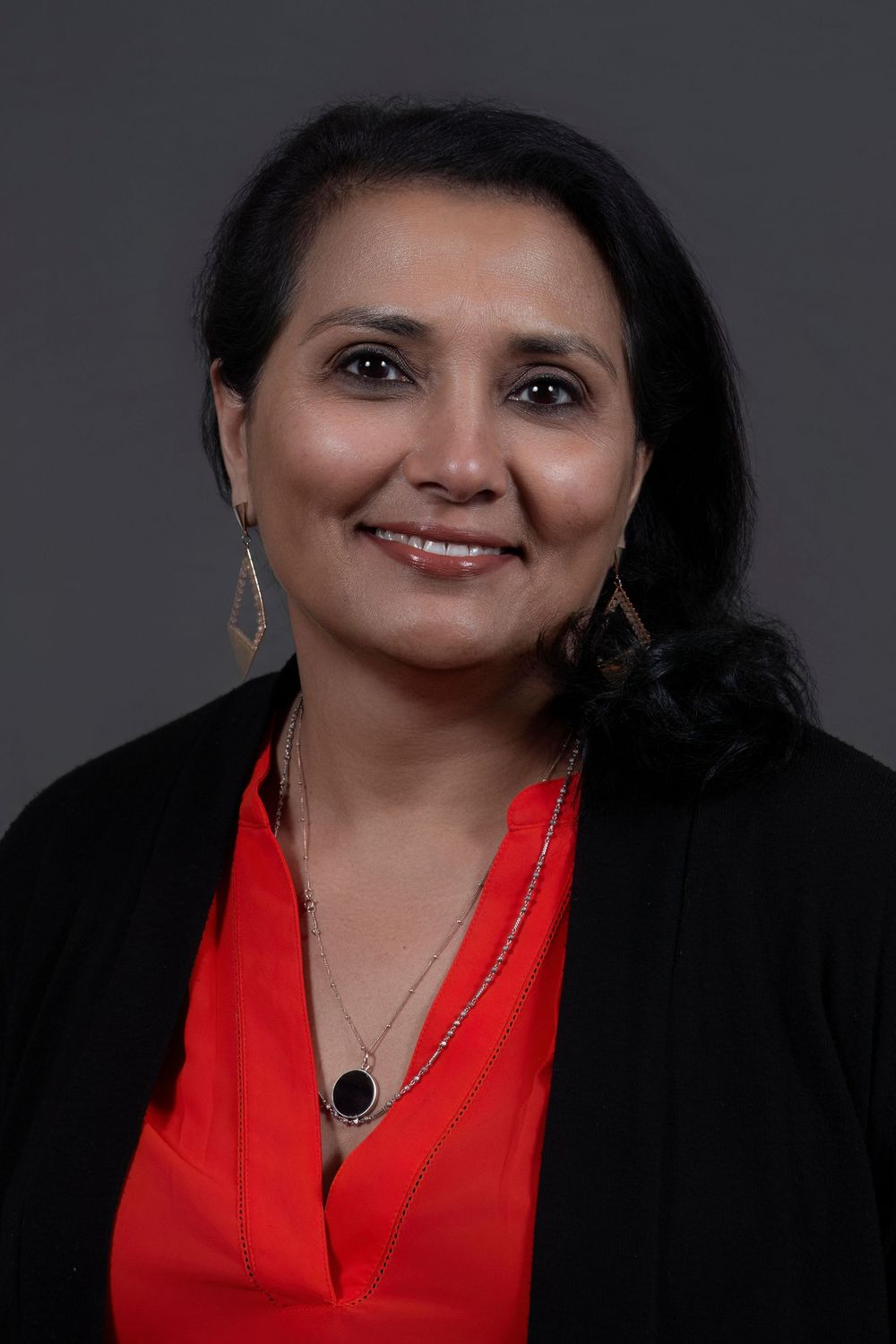 Professional headshot of a person wearing a black blazer and red blouse with dangling earrings against a dark background. In Orlando Fl