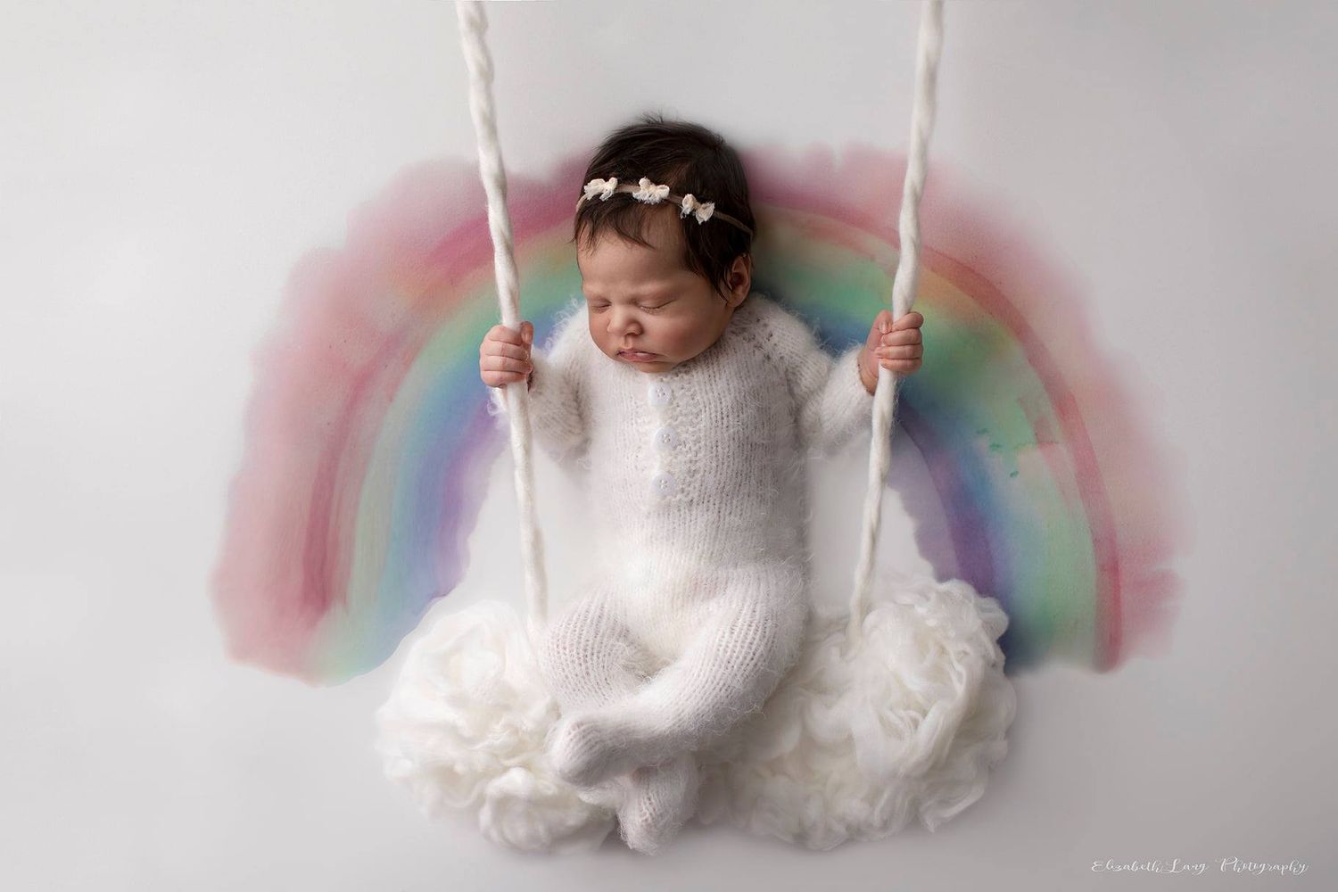 A sleeping baby in white clothing lies on a rainbow cloud swing against a light gray background.