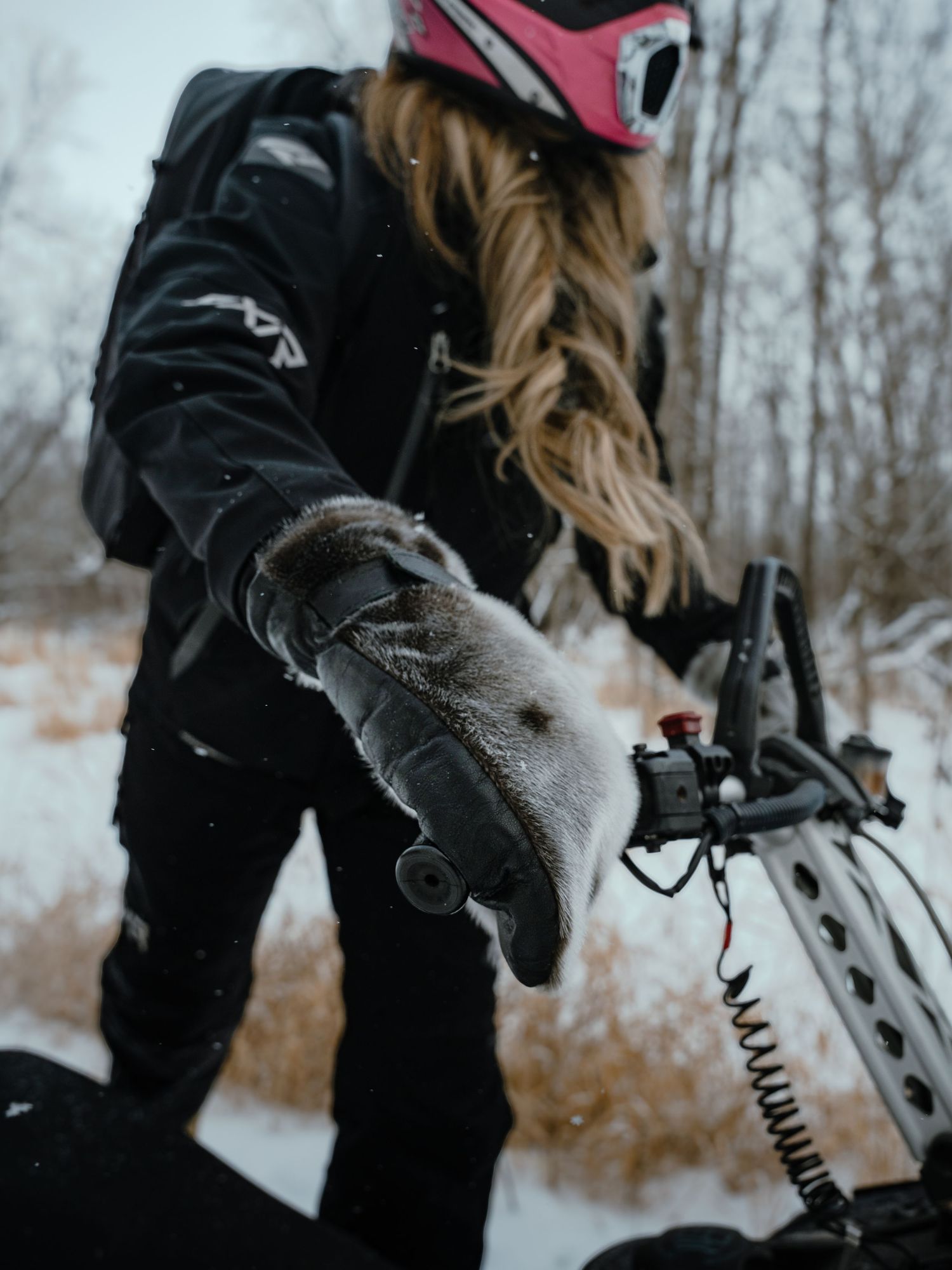 Winter sports enthusiast in black gear adjusts equipment on snowy terrain with trees in background.