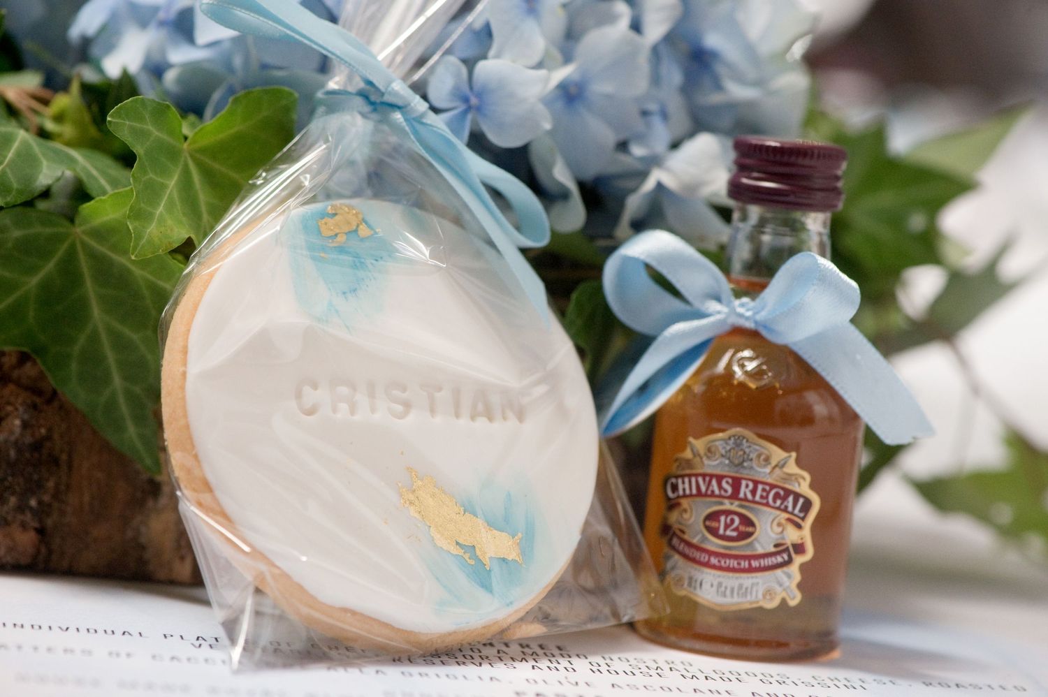 Personalized Cristian cookie favor and mini Chivas Regal whisky bottle with blue ribbon bow on floral background.