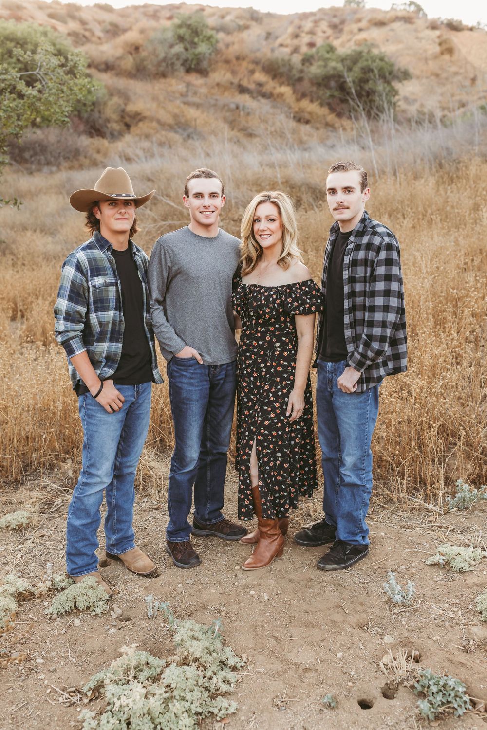A group in western attire poses together on a dusty hillside wearing plaid shirts, jeans and cowboy boots.