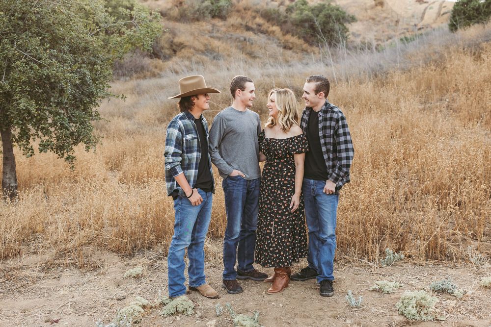 A group of four people stand together on a hillside wearing casual western attire against a golden dry grass backdrop.
