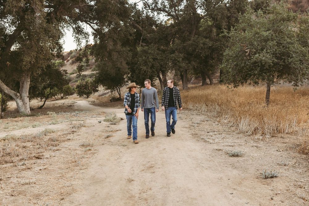 Three people walk down a dirt path through dry grass and trees on a sunny day.