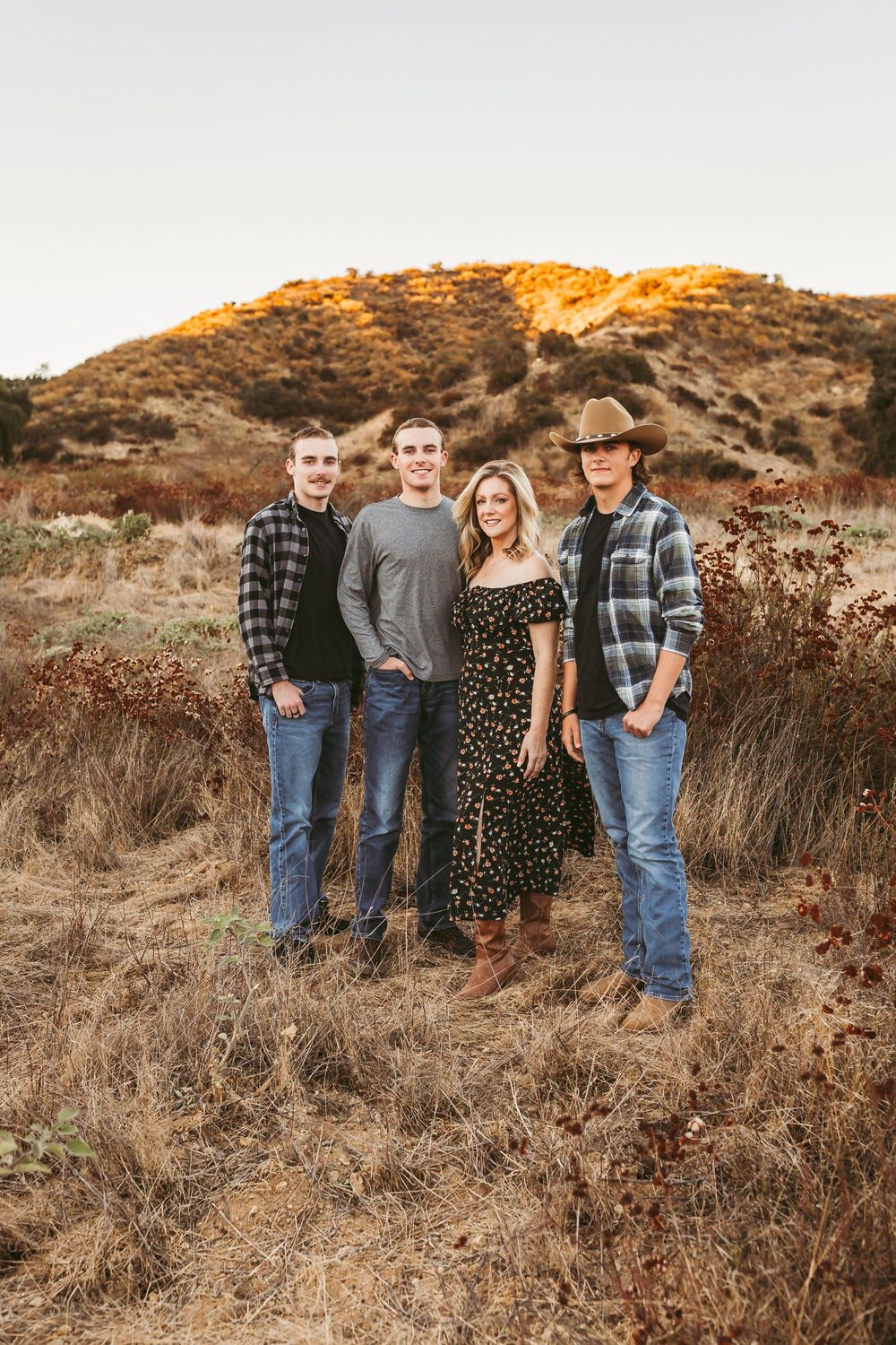 A casual outdoor portrait session at golden hour with people in western wear against a rustic hillside backdrop.