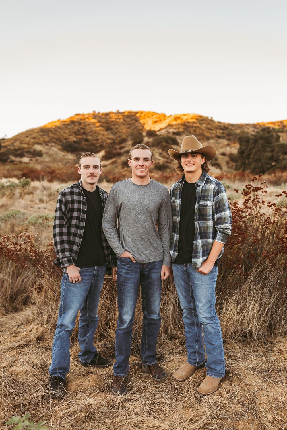 Three people in casual western wear stand together in a field during golden hour sunset.