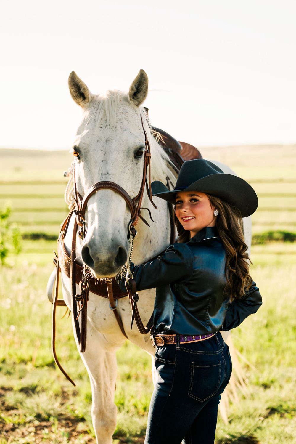 A white horse stands in a grassy field while someone in western attire interacts with it.