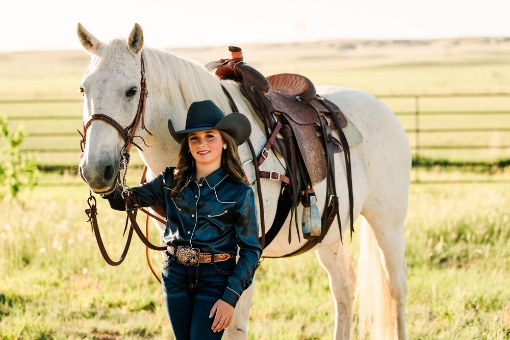 A western rider poses with a white horse wearing traditional riding gear and saddle in a grassy field.