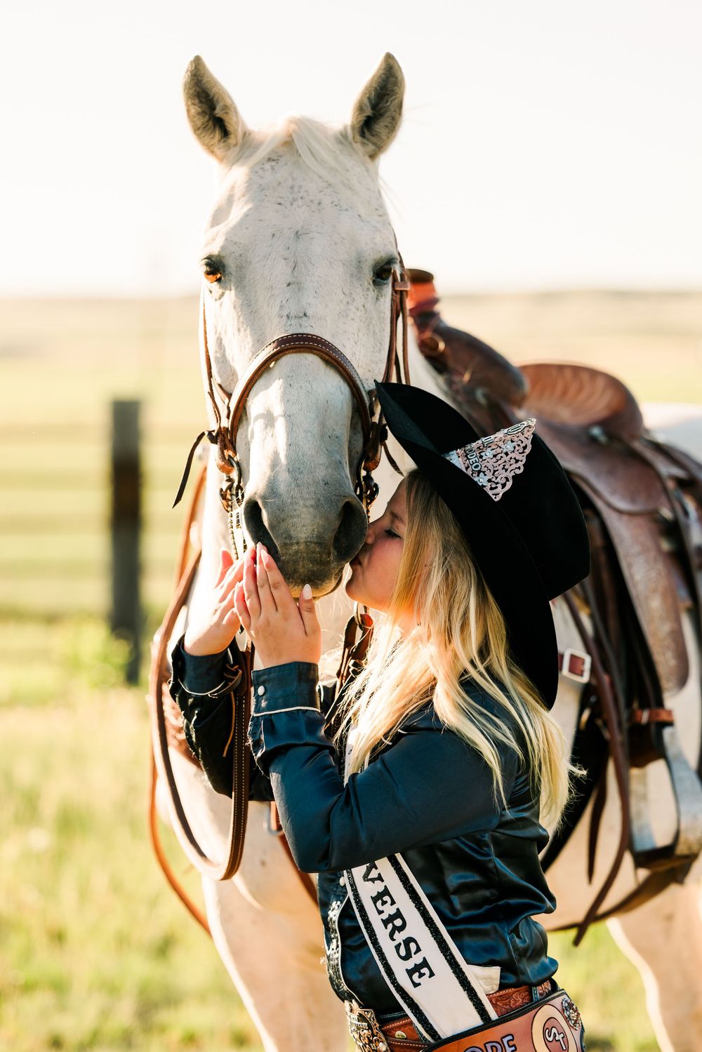 Close-up of white horse with western tack and rider in navy jacket against pastoral background.