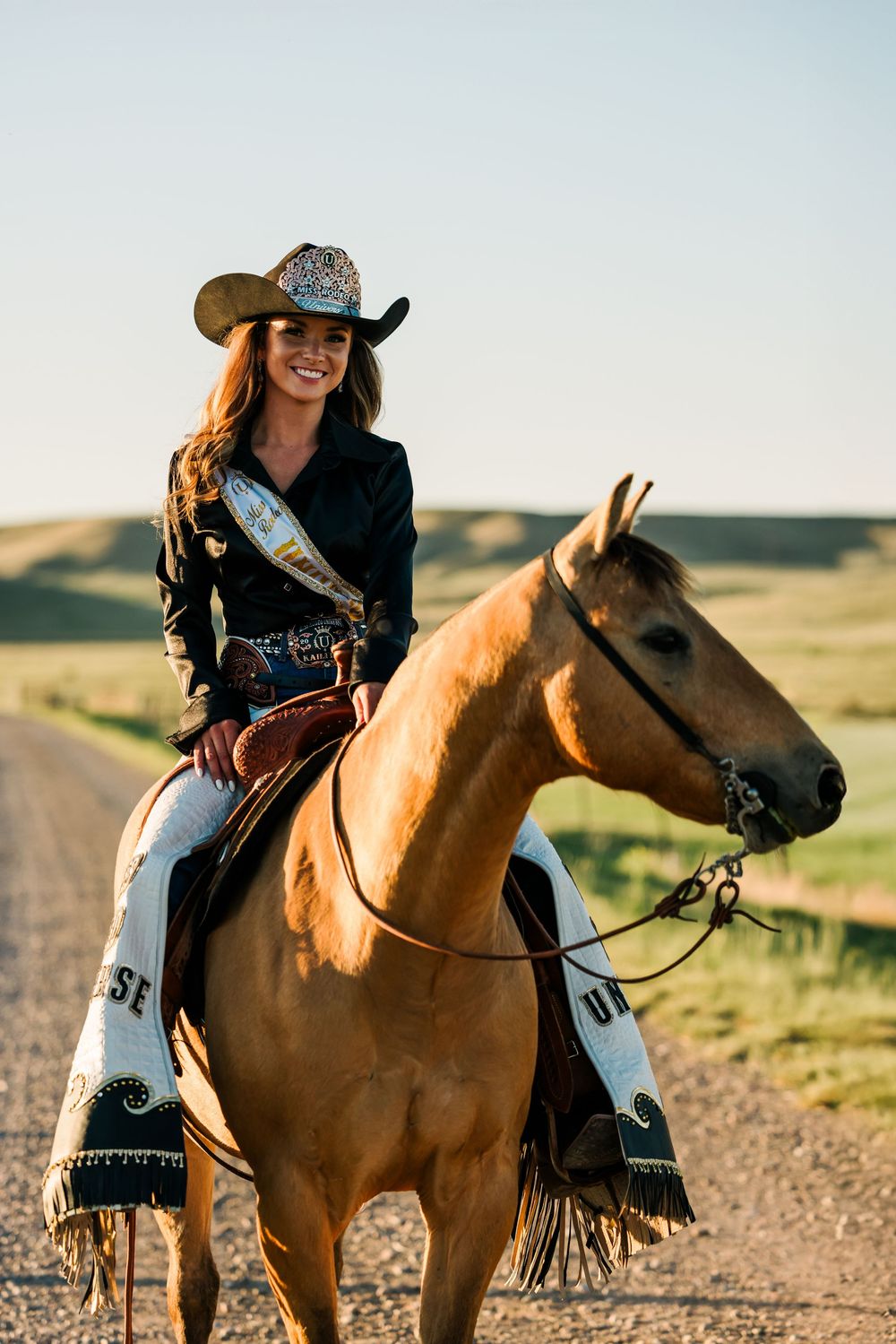 A rider in western attire sits atop a chestnut horse on a dirt road with rolling hills in the background.