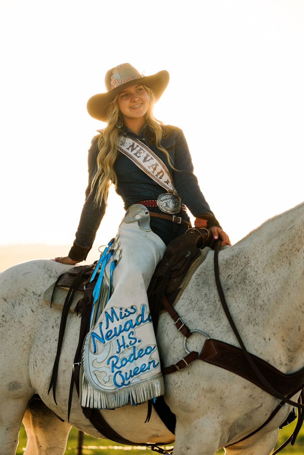 Rider wearing rodeo queen sash sits on decorated western horse during golden hour.