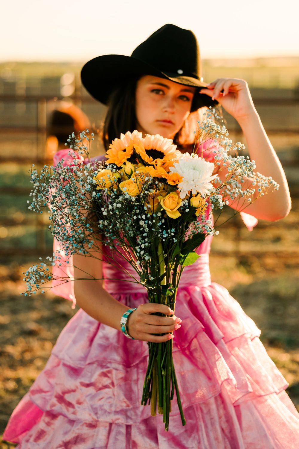 A figure in a pink ruffled dress and black cowboy hat holds a bouquet of yellow and white flowers at sunset.