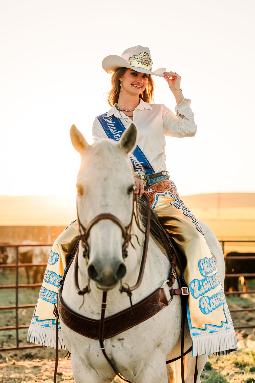 Western rider in white fringe outfit poses on white horse against sunset backdrop.