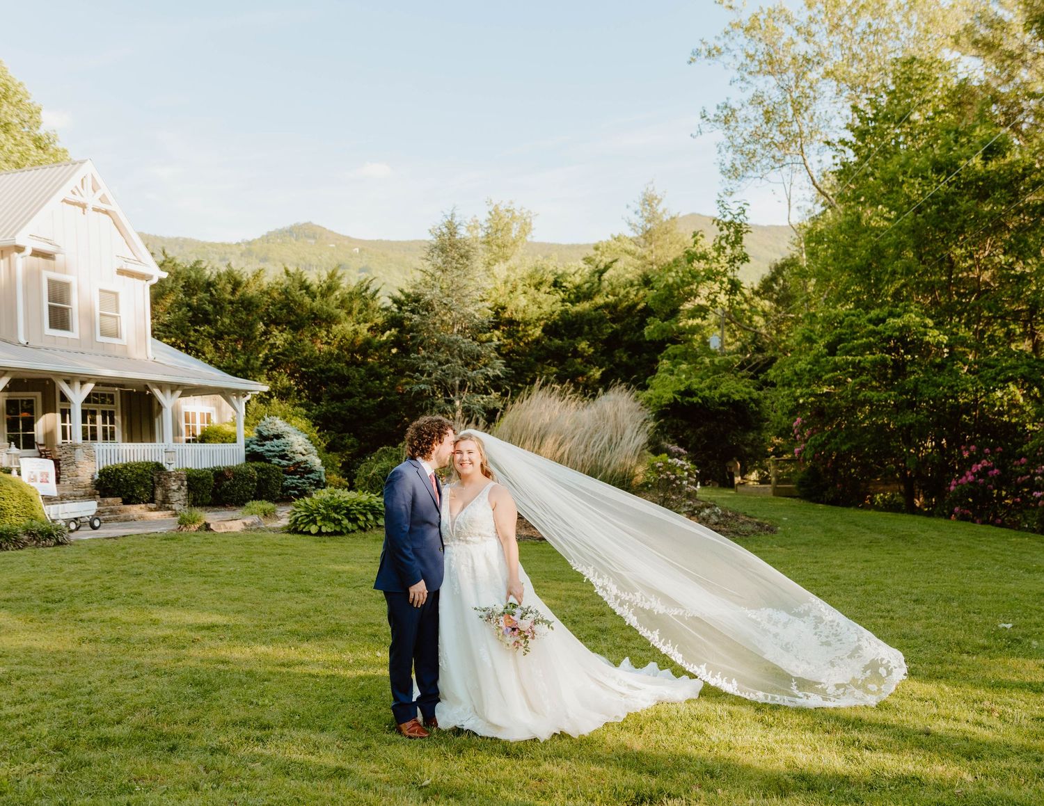 Wedding couple poses in front of a white farmhouse on a scenic grassy hill.