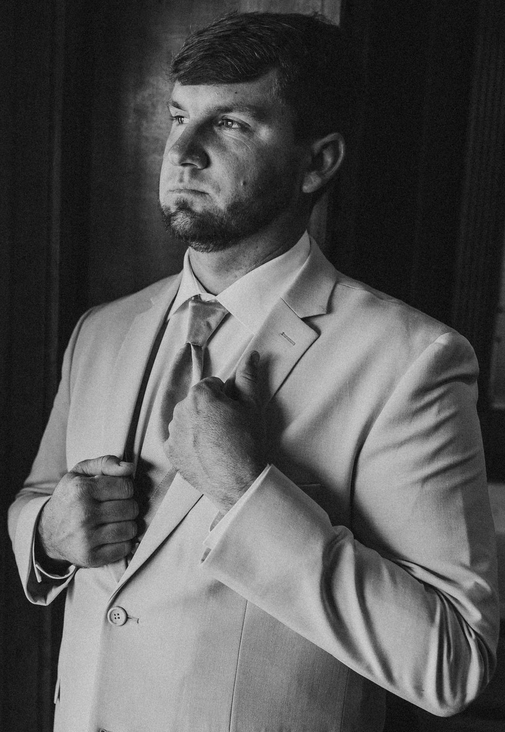 Black and white photo of a man in a light suit adjusting his tie, looking away dramatically.