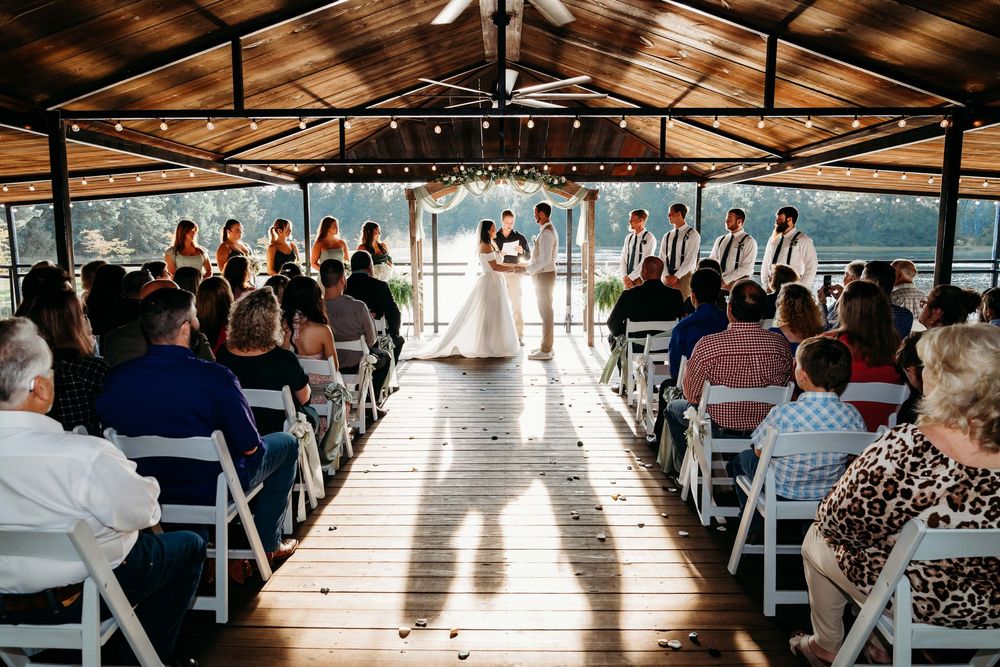Wedding guests seated in white chairs under a covered pavilion during an outdoor ceremony with sunlight streaming in.