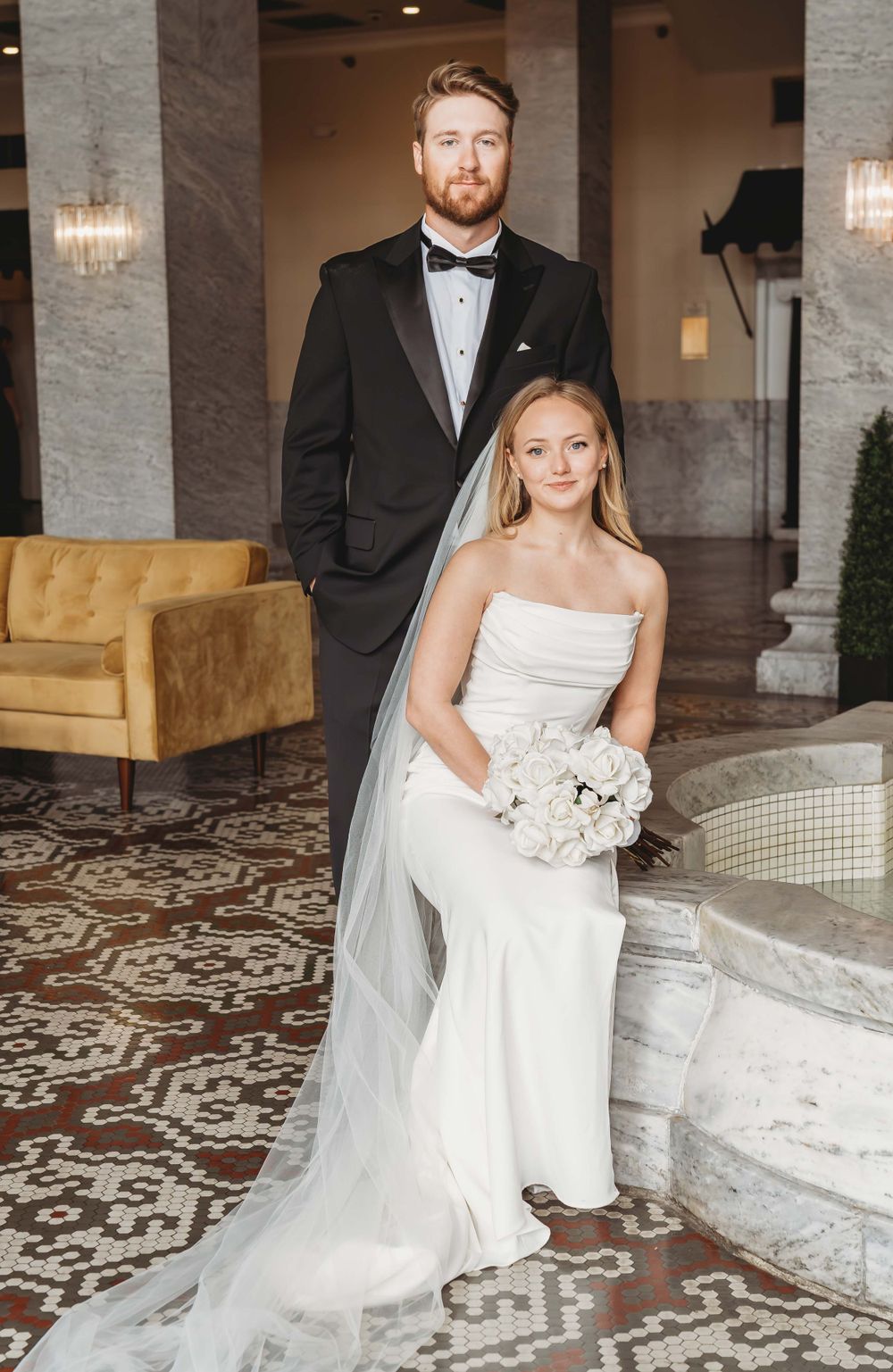 A bride in a fitted white gown and long veil poses with a groomsman in black tuxedo at an elegant venue.