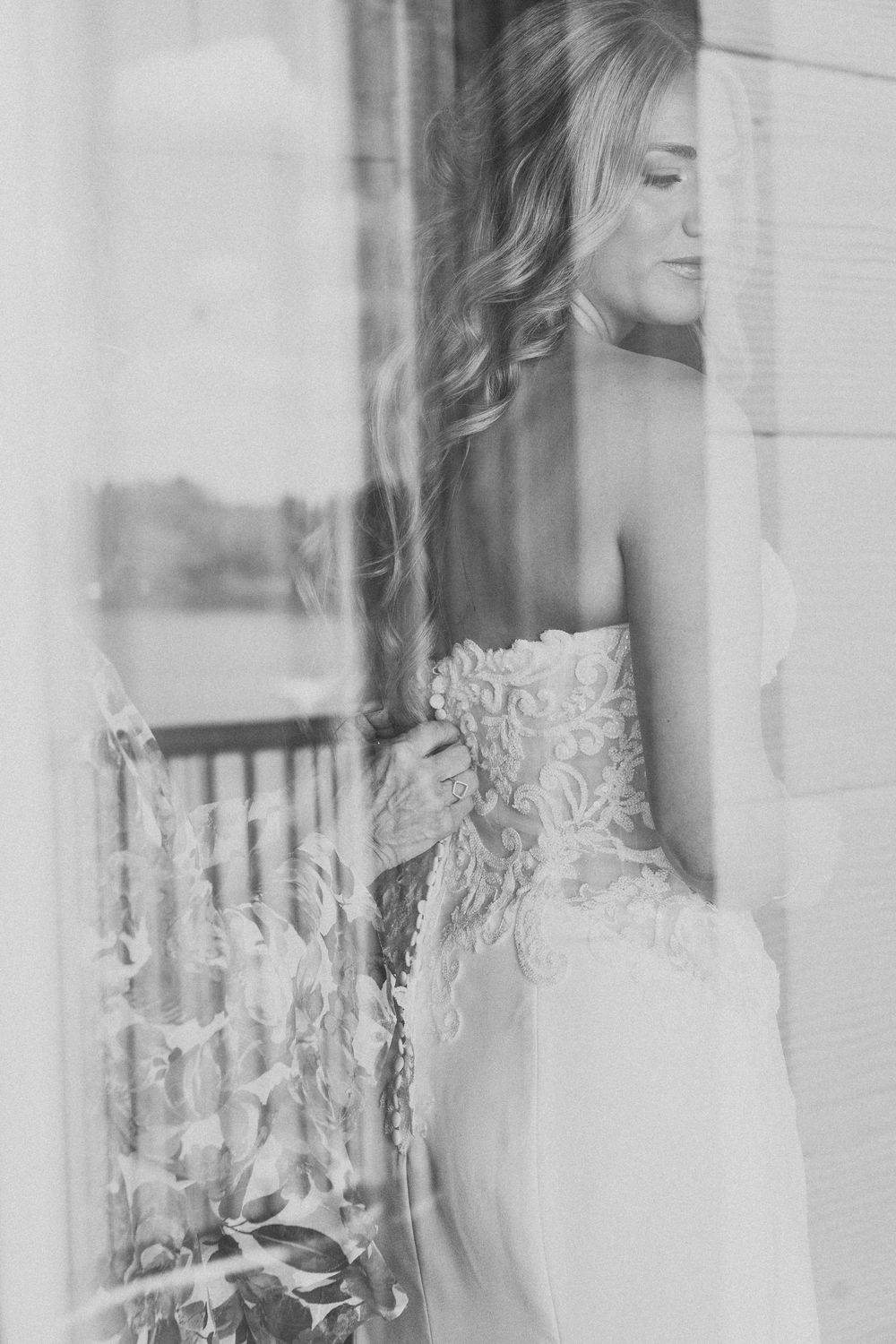 Black and white photo of a bride in a lace strapless gown gazing out a window on her wedding day.