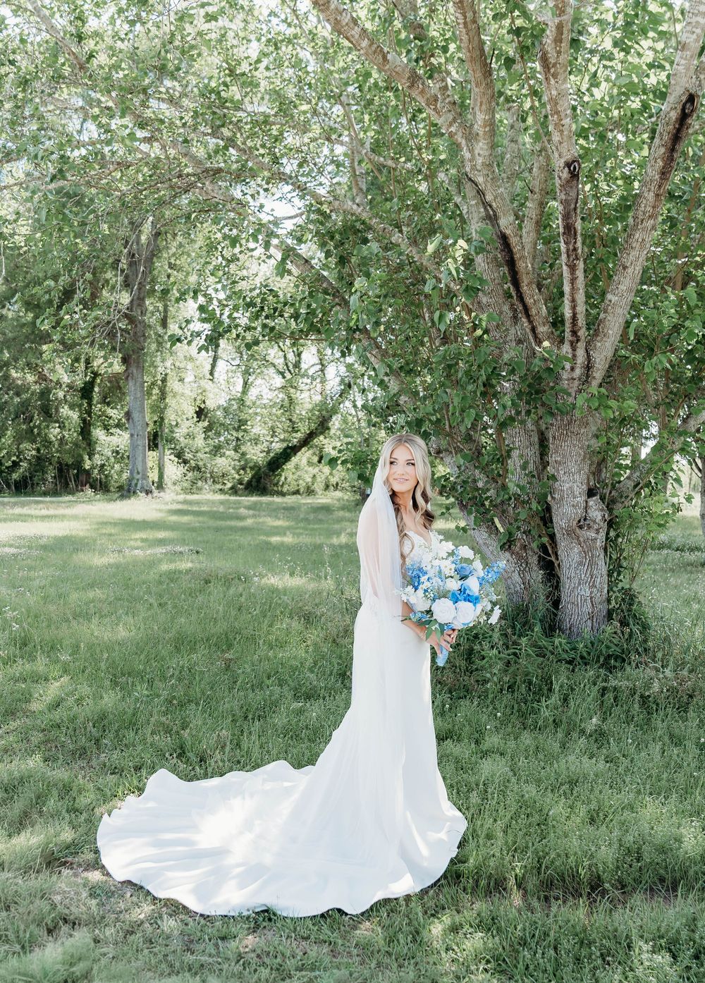 Bride in white gown with train holds blue bouquet beside large tree in lush green outdoor setting.