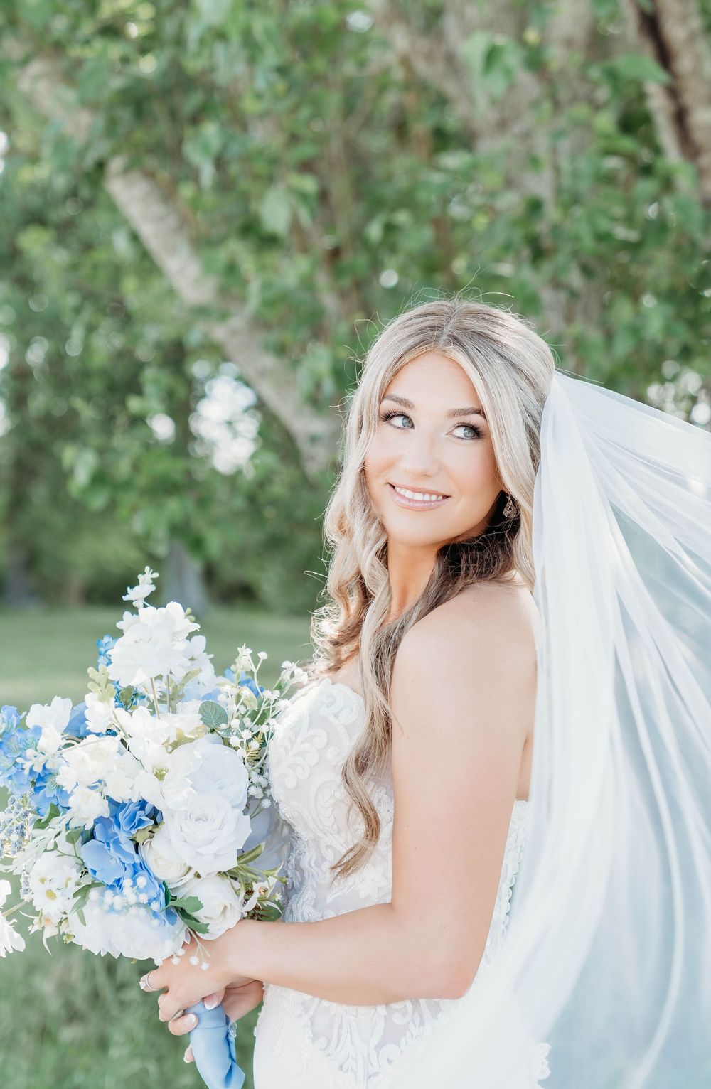 Smiling bride in a lace wedding dress holding a blue and white floral bouquet with a flowing veil outdoors.