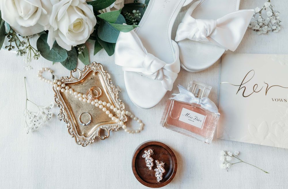 Elegant wedding styling flatlay with white roses, perfume bottles, jewelry, and eucalyptus on light beige background.