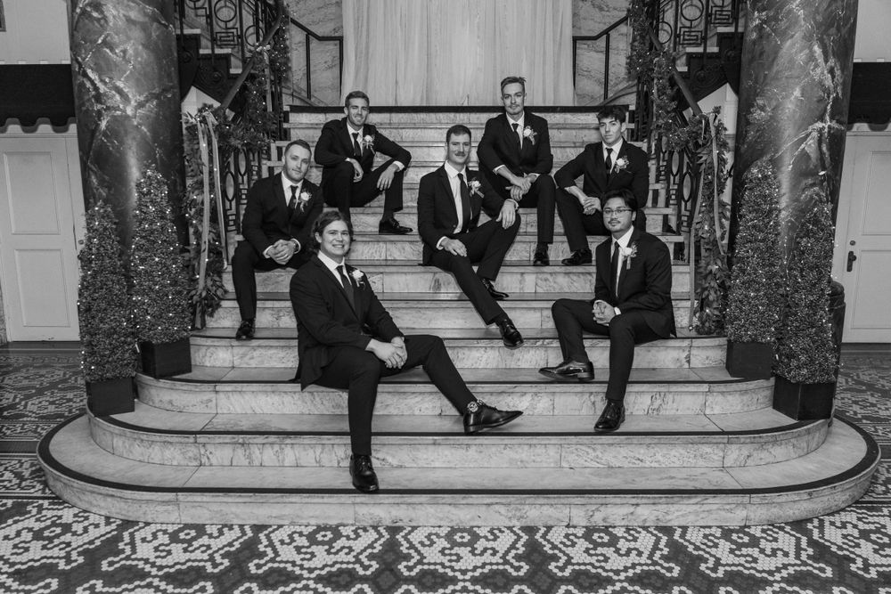 Group of groomsmen in black tuxedos posed formally on ornate marble steps.