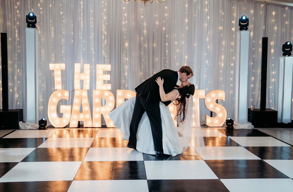 Couple shares romantic dance dip on black and white checkered floor with illuminated marquee letters in background.