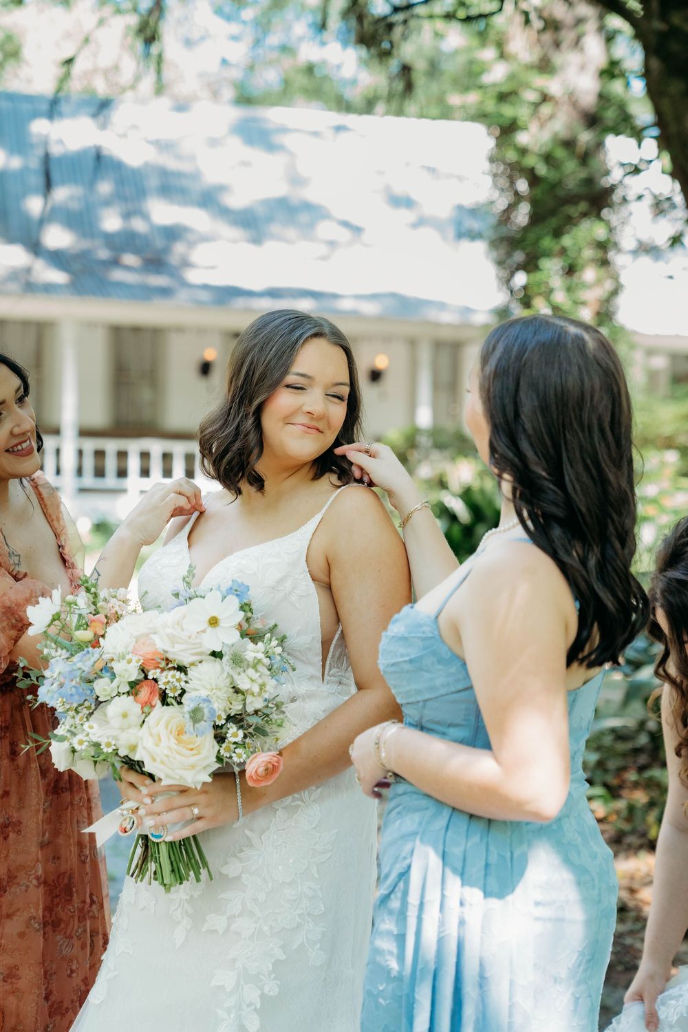 Bride in white lace gown holds pastel bouquet while bridesmaid in blue adjusts her hair outdoors.