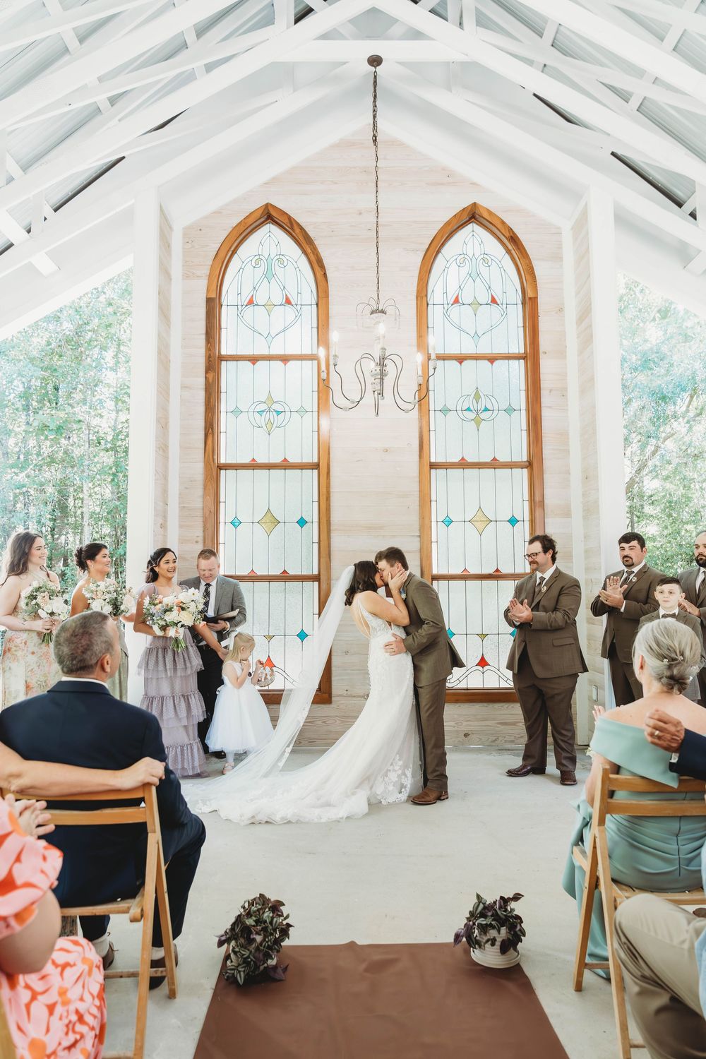 Bride and groom share first kiss at altar in white chapel with stained glass windows surrounded by wedding party and guests.