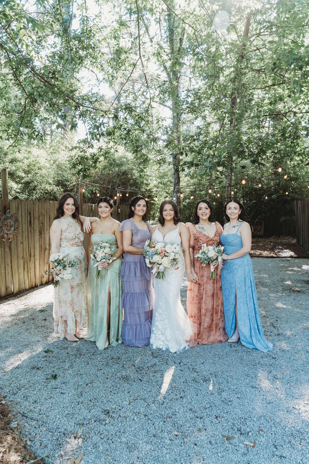 Bride in white lace gown poses with five bridesmaids in colorful mismatched dresses holding bouquets outdoors.