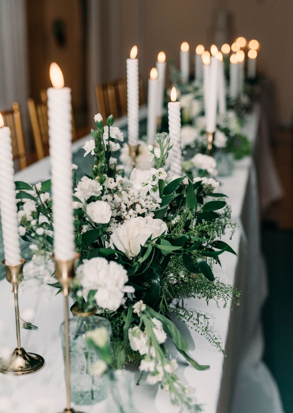 Elegant wedding table centerpieces featuring white candles, white roses and greenery in glass vases create a romantic atmosphere.