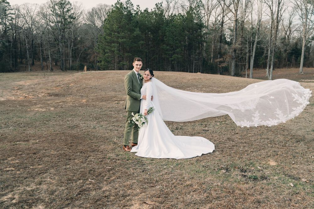 Long flowing white wedding dress train spread across outdoor field with trees.