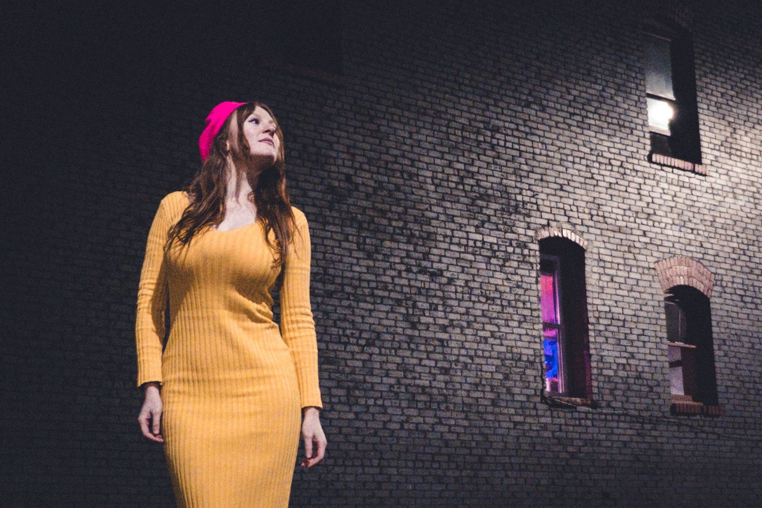 A mustard yellow sweater dress stands against a dark brick wall with illuminated windows.