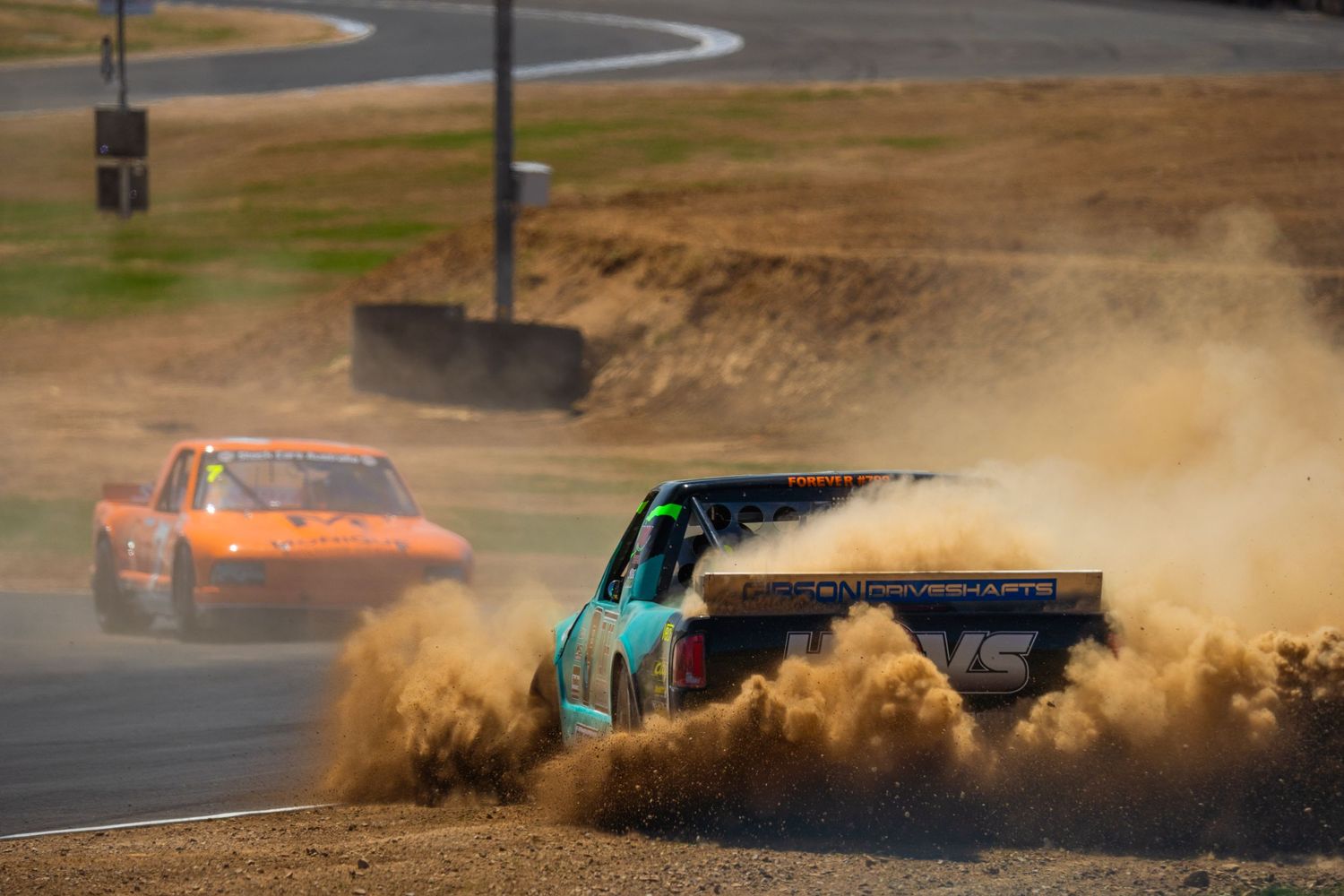 Rally cars kicking up dirt and dust while racing around dirt track corner.