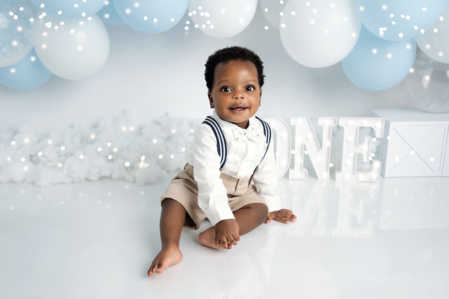 Baby in white outfit sitting against blue and white balloon backdrop.