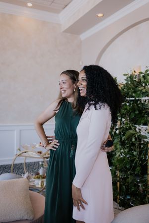 Two friends pose together at an elegant indoor event, one wearing a forest green dress and one in blush pink.